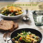 Two plates of green bean soup on a tablecloth with glasses and a slice of bread.